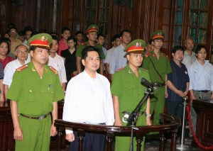 Cu Huy Ha Vu stands between policemen in the dock during his trial at a court in Hanoi